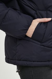 Veste matelassée bleu marine avec texture en losanges, dotée d'une poche latérale et d'un cordon de serrage réglable à l'ourlet. Matériau doux et léger.