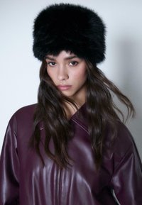 Young woman with long dark hair wears a black fur hat and a dark purple leather jacket, facing forward against a plain background.