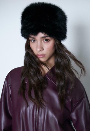 Una giovane donna con lunghi capelli scuri indossa un cappello di pelliccia nera e una giacca di pelle viola scuro, rivolta in avanti su uno sfondo semplice.