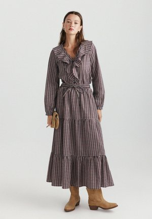 Femme portant une robe midi à manches longues à carreaux avec des volants, une taille nouée, des bottines beige et tenant une petite pochette beige.