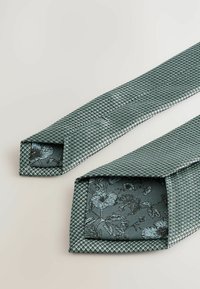 Lys tealfarvet slips med et struktureret diamantmønster. Indersiden har et blomstermønster i mørkere nuancer. Kanterne er kvadratskåret.