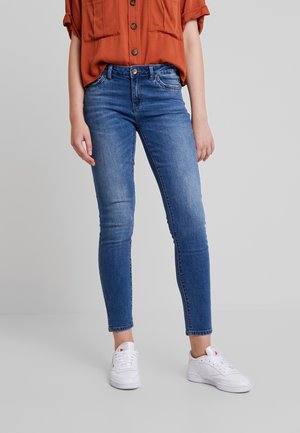 Osoba ubrana w niebieskie obcisłe dżinsy, białe trampki oraz rdzawą koszulę zapinaną na guziki z podwiniętymi rękawami na jednolitym szarym tle.