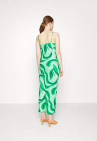 Vestido verde com padrão de folhas onduladas, alças finas, ajuste solto e comprimento longo. Usado com sandálias de salto laranja.