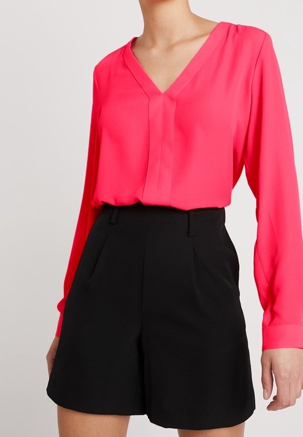 Femme portant un chemisier rose vif à manches longues rentré dans un short noir taille haute, sur un fond uni.