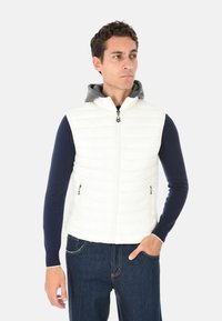 Uomo che indossa un gilet bianco trapuntato senza maniche sopra un maglione blu navy e jeans scuri, rivolto leggermente a destra con una mano in tasca.