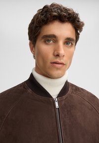 Bruine suède bomberjack met een ribgebreide kraag en ritssluiting, gedragen over een licht crème turtleneck trui met een gestructureerde stof.