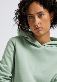 Felpa verde menta in tessuto morbido e testurizzato, con cappuccio regolabile e vestibilità comoda. Cuciture evidenti e stile informale.
