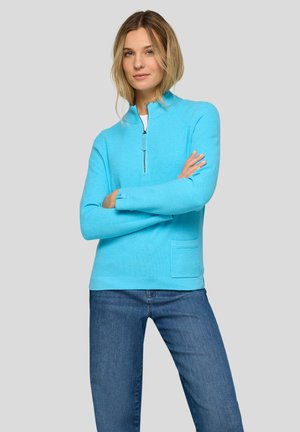 Frau mit blondem Haar, die einen leuchtend blauen Pullover mit Viertelreißverschluss und eine blaue Jeans trägt, steht mit verschränkten Armen vor einem grauen Hintergrund.