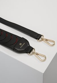 Czarny skórzany pasek torebki z złotymi akcentami, wytłoczonym logo, teksturowaną powierzchnią i okrągłymi czerwonymi detalami z jednej strony.