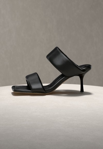 Μαύρο δερμάτινο mule με ψηλό τακούνι, δύο φαρδιές λουρίδες και τετράγωνη μύτη, τοποθετημένο σε φωτεινή επιφάνεια με ουδέτερο φόντο.
