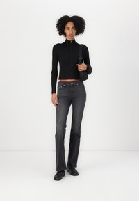 Black ribbed zip-up top with red trim, paired with dark gray flared jeans. Model wears black shoes and holds a black strap bag.