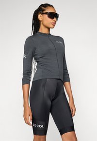 Sorte sykkelshorts med polstret sete og hvit logo, kombinert med en grå langermet topp med glidelås og tettsittende design.