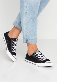 Zapatillas de lona negras con suelas y cordones de goma blanca, que presentan un diseño clásico de corte bajo y un capuchón blanco en la punta. Pantalones de mezclilla con los puños remangados son visibles.