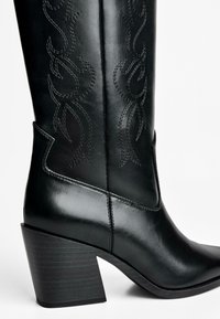 Black leather cowboy boot with a pointed toe, ornate stitching along the shaft, and a chunky wooden heel. Smooth texture, modern design.