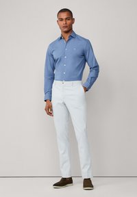 Camisa de botones azul claro, pantalones blancos de corte slim y zapatos oscuros de slip-on. Tela suave, corte ajustado y diseño minimalista.