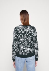 Maglione verde scuro con motivo di fiocchi di neve bianchi, tessuto a maglia, maniche lunghe, orlo a coste, indossato sopra un paio di jeans azzurri.