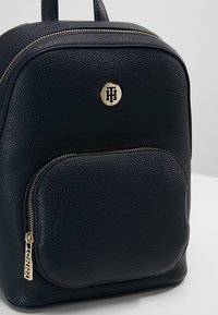Sac à dos en cuir noir texturé avec poche zippée à l'avant, fermetures éclair dorées, et un emblème rond doré avec des initiales sur la partie haute avant.