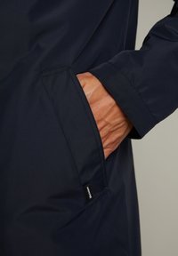 Chaqueta impermeable azul marino con un bolsillo lateral, textura suave y etiqueta de marca sutil en la costura. Primer plano de la tela y una mano.