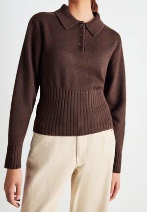 Vrouw draagt een bruin gebreide polo met lange mouwen, geribde manchetten en tailleband, gecombineerd met lichtbeige broek.