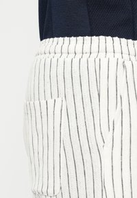 Pantalon de détente rayé blanc avec des rayures foncées, doté d'une taille à cordon de serrage et d'une poche latérale. Le tissu semble doux et texturé.