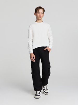 Camicia bianca a maniche lunghe abbinata a pantaloni cargo neri con grandi tasche laterali. Il look è completato da sneakers bianche e nere.