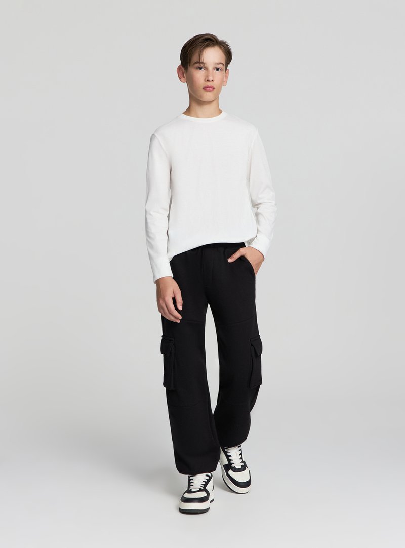 Biała koszula z długim rękawem w połączeniu z czarnymi spodniami cargo z dużymi bocznymi kieszeniami. Całość dopełniają czarno-białe trampki.