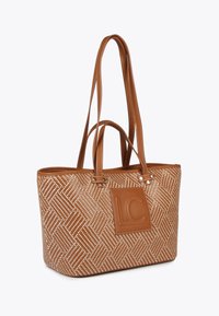 Tan woven tote bag with cream diagonal pattern and tassel detail. Features two long handles and a side logo patch. Made of textured material.