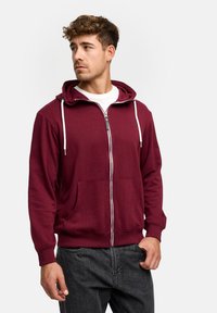 Burgundy zip-up hoodie made of soft fabric, featuring a front pocket, white drawstrings, and ribbed cuffs and hem. Styled with a white shirt underneath.