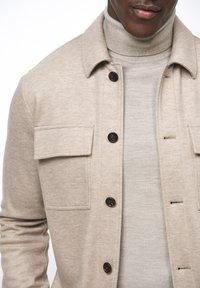 Beige button-up jack met borstzakken, gemaakt van een textuurstof. Gdragen over een lichtbeige coltrui. Kenmerkt zich door donkere knopen.