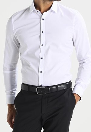 Homme portant une chemise blanche ajustée à boutons noirs, un pantalon noir et une ceinture noire, les mains posées négligemment dans les poches.