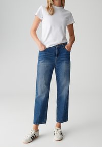 Femme portant un t-shirt blanc uni à manches courtes, un jean bleu droit et des baskets blanches avec des rayures noires, debout les mains dans les poches.