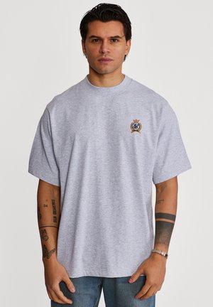 Uomo con capelli scuri che indossa una maglietta oversize grigio chiaro con un piccolo logo ricamato sul petto, jeans blu e tatuaggi visibili sulle braccia.