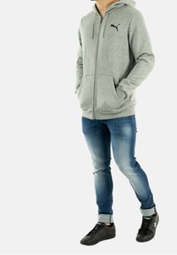 Gray zip-up hoodie with kangaroo pockets, black logo, worn with blue jeans and black sneakers, featuring rolled cuffs and a slim fit.