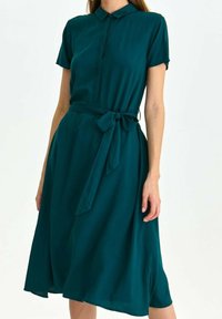 Tygklänning i teal med korta ärmar, knappstängd framtill och knytbart midjeskärp. Flödande tyg med knälång, A-linjeformad silhuett.