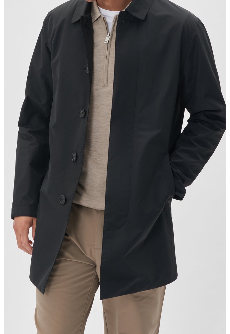Black water-resistant coat with a straight cut, button front, and flat collar. Lightweight fabric, side pockets, worn over a beige top.