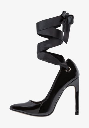 Chaussure à talon haut en cuir verni noir avec un bout pointu et des lacets en ruban autour de la cheville, comportant un œillet pour le ruban au niveau de la cheville.