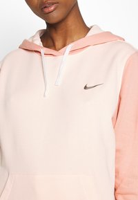 Person som bär en ljusrosa och persikofärgad Nike-hoodie med en vit dragsko och framficka, avskuren under näsan och ovanför midjan.