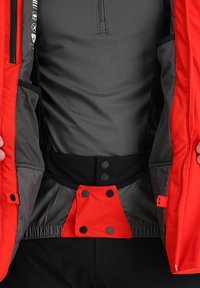 Rote isolierte Jacke mit grauem Innenfutter, ausgestattet mit mehreren Innentaschen, Druckknöpfen und Reißverschlüssen. Weiche Textur mit elastischem Taillenbund.