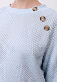 Maglione azzurro chiaro con texture, caratterizzato da motivi a coste diagonali e tre bottoni decorativi marroni sulla spalla. Tessuto morbido, vestibilità rilassata.