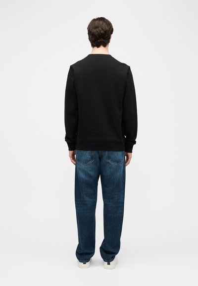 Άνδρας που φοράει μαύρο μακρυμάνικο πουκάμισο, φαρδιά μπλε τζιν με πίσω τσέπες και λευκά αθλητικά παπούτσια, στέκεται αντικρίζοντας ένα απλό λευκό φόντο.