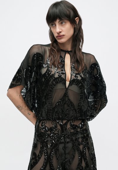 Hosszú sötét hajú nő fekete, áttetsző ruhát visel, amelyen flitteres virágminták és mély kivágás található, és egy egyszínű világos háttér előtt áll.
