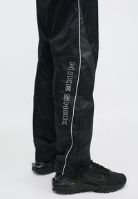 Mustad mustaharjoituspüksid, millel on sile pind, tumedad läikivad paneelid ja valged õmblused. "NIKESHOX" logo on vertikaalselt küljele trükitud.