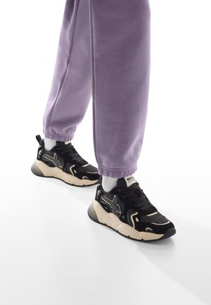 Sneaker nere e beige con upper testurizzato, dettagli riflettenti e suola spessa. Abbinate a pantaloni leisure di colore viola chiaro con polsini elasticizzati.