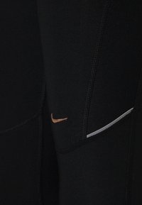 Czarne legginsy sportowe wykonane z elastycznego materiału, z płaskimi szwami, małym miedzianym logo Nike oraz białym akcentem po boku.