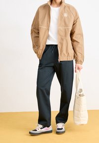 Light brown zip-up jacket with white accents, paired with dark pants and multicolored sneakers. Beige tote bag in hand.
