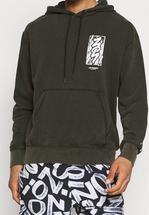 Mężczyzna w ciemnozielonej bluzie z kapturem z białym nadrukiem i czarno-białych wzorzystych spodniach z pogrubionymi literami.