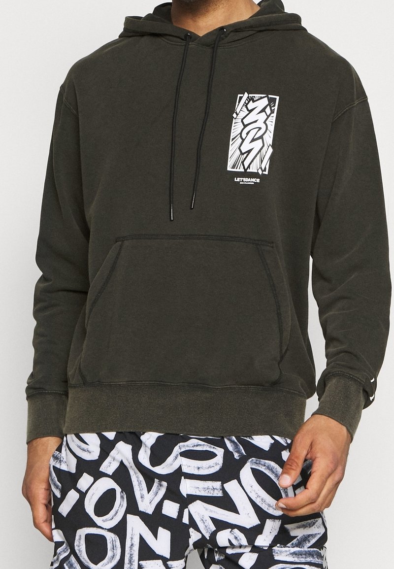 Homme portant un sweat-shirt à capuche vert foncé avec un motif graphique blanc et un pantalon à motifs noir et blanc avec des lettres en gras.