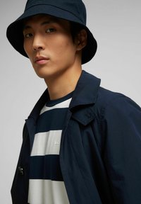 Navy blue trench coat with a pointed collar, worn over a white and navy striped shirt. A navy bucket hat completes the look.