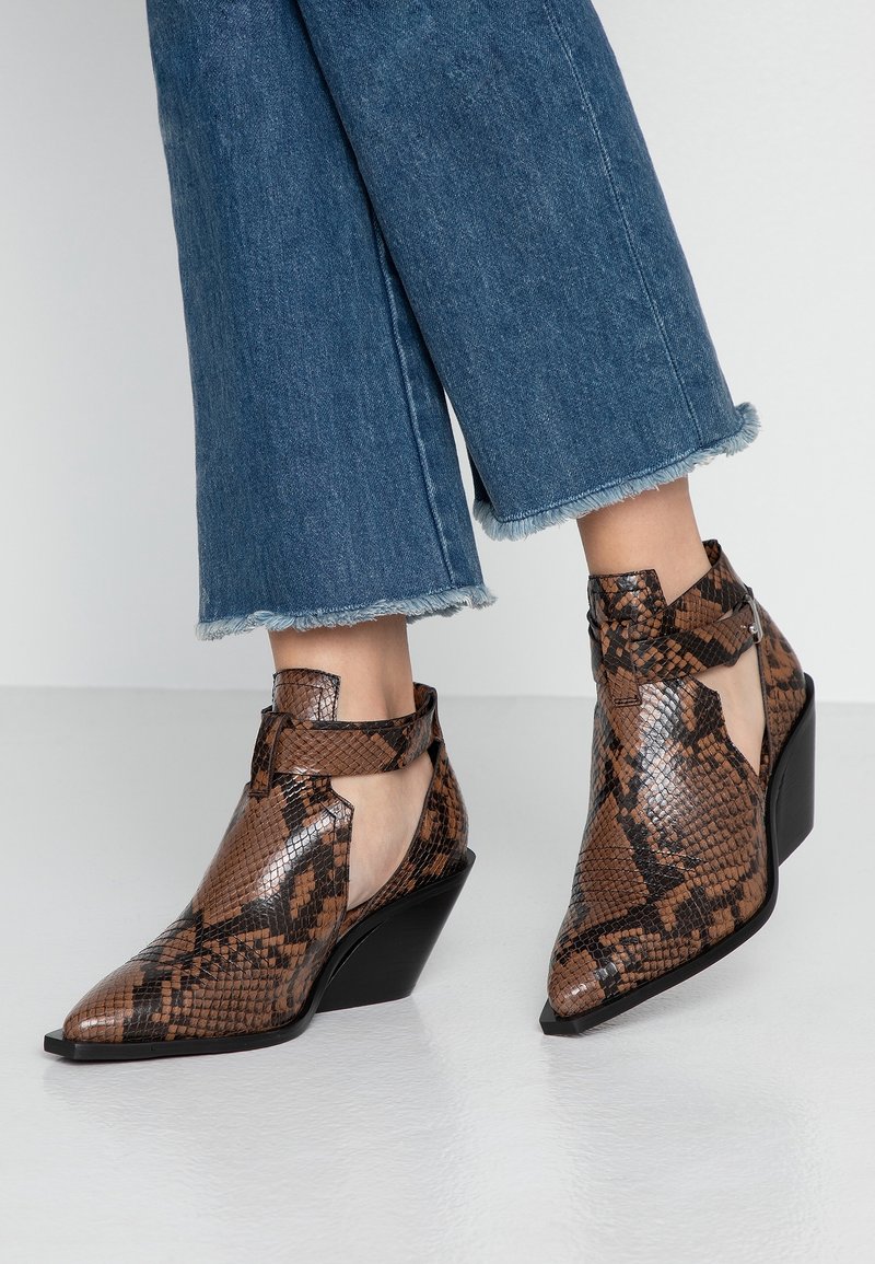 Bottes cheville à imprimé serpent marron avec une pointe effilée, présentant un design découpé et un talon compensé bas, finies avec une lanière à la cheville.