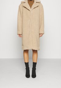 Beiger Kunstpelzmantel mit Kragen, langen Ärmeln und geradem Schnitt, der bis zur Mitte der Oberschenkel reicht. Getragen über einem gestreiften Oberteil und schwarzen Socken.